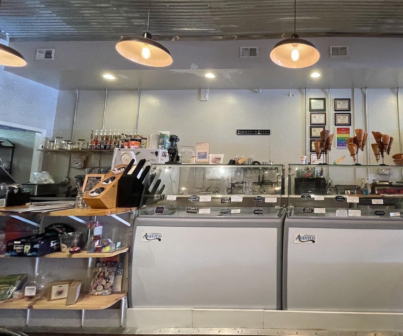 The service counter at Magnolia Market: two ice cream cases with hand written labels, waffle cones on stands, a glass jar of muffins, stacked pizza boxes, an oven and coffee equipment under pendant lights.