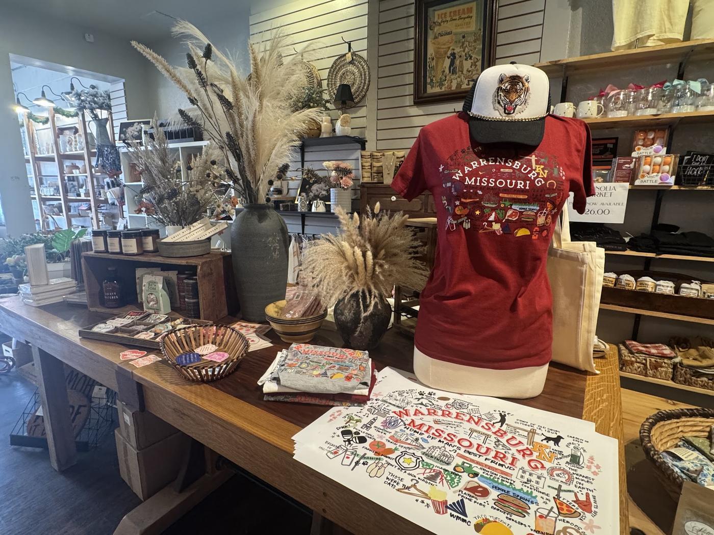 The shop floor at Magnolia Market: Warrensburg Missouri merchandise on a wood table, dried grasses in stoneware, candles and jars, and shelves of mugs and goods behind.